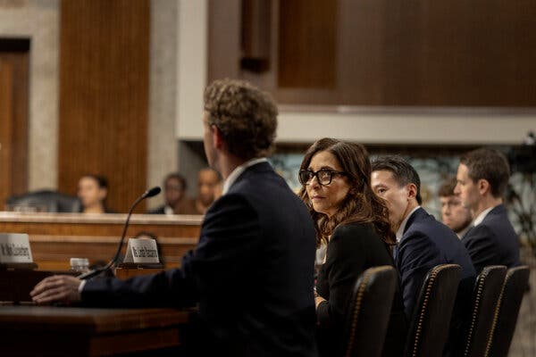 Linda Yaccarino sits at a table in a hearing room. People sit on either side of her.