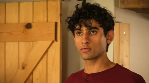 BBC/Moonage Pictures A young man is stood in a house, looking slightly away from the camera