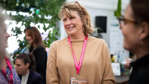 Cheltenham Literature Festival Nicola Tuxworth, in focus, smiling to someone off camera, wearing a beige jumper and a pink lanyard, with dusty blonde hair and glasses on top of her head, surrounded by people out of focus