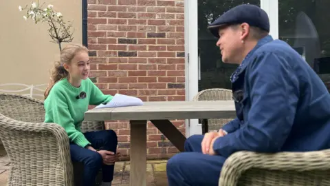 Jon Wright/BBC Anna, an 11 year old girl, wears a green jumper and sits at a garden table with poet Luke Wright