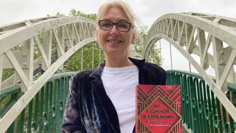 Alex Pope/BBC Ruth Hogan standing on The Suspension Bridge in Bedford holding her book